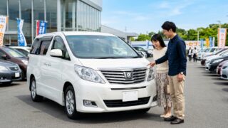 中古車展示場に並ぶパールホワイトの20系アルファードの前に立ち笑顔で車両状態を確認する若い日本人夫婦