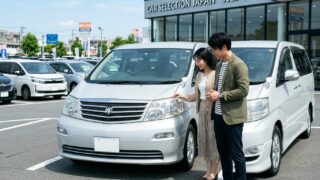 中古車販売店の展示場に並ぶ初代10系アルファードの前に立ち車両状態とプライスボードを真剣に確認する若い日本人夫婦