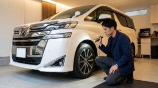 明るい日本の自宅ガレージでパールホワイトのヴェルファイアのフロントタイヤと足回り周辺をしゃがんで点検する若い日本人男性