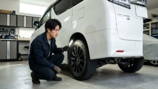 明るい日本の自宅ガレージでパールホワイトのヴェルファイアのリアタイヤ内側の摩耗状態をしゃがんで点検する若い日本人男性